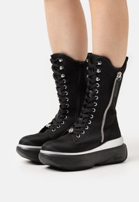 Botas altas negras con cordones, acabado mate suave, detalles en hardware plateado y suela gruesa blanca. Incluye cierre lateral.