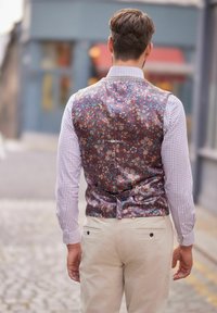 Brązowa kamizelka w kwiatowy wzór o gładkiej fakturze, noszona na kraciastej koszuli, zestawiona z jasnobeżowymi sztruksowymi spodniami.