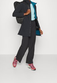 Chaqueta negra impermeable con forro turquesa, combinada con una camisa azul clara, pantalones oscuros y zapatillas deportivas rosas. Mochila llevada en la espalda.