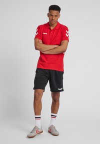 Rød polo skjorte med korte ærmer og hvidt logo, parret med sorte shorts med logo. Hvide strømper med røde og sorte striber, grå sko.