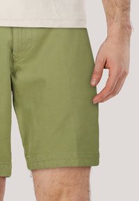 Pantaloni verdi realizzati in tessuto testurizzato, con un design tradizionale e orlo piegato, mostrati con una mano poggiata sul fianco.