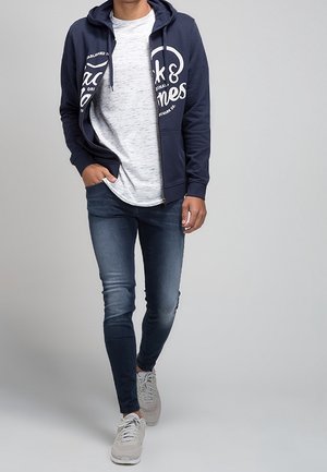 Sweat à capuche zippé bleu marine avec un grand motif blanc, associé à un t-shirt gris clair et un jean skinny bleu foncé. Des baskets claires complètent la tenue.