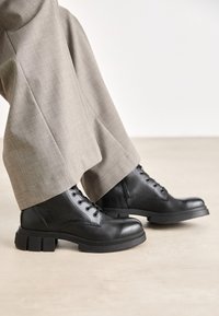 Bottines en cuir noir avec un design à lacets, semelle en caoutchouc épaisse et bout rond, associées à un pantalon ajusté gris clair.