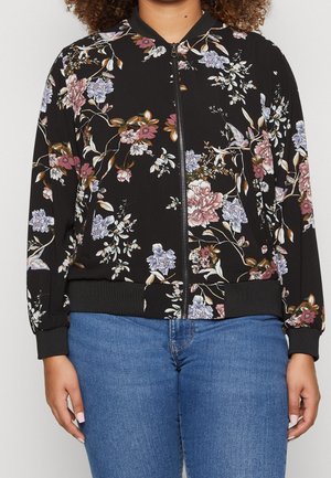 Zwarte bomberjas met ritssluiting, geribde manchetten en zoom, met een bloemenpatroon in roze, paars, wit en groen, gedragen met blauwe jeans.