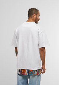 Un bărbat cu părul scurt stă cu spatele, purtând un tricou simplu, alb, supradimensionat, peste o cămașă cu model colorat și blugi albastru deschis.