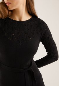 Svart stickad tröja med långa ärmar som har ett strukturerat diamantmönster och ett bälte i midjan. Slät tyg med rundad halsringning.