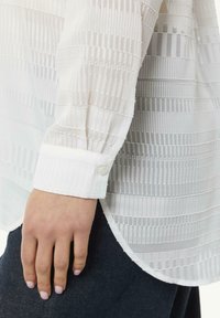 Weiße Bluse mit langen Ärmeln, die über strukturierte Streifen, eine Knopfmanschette und einen abgerundeten Saum verfügt. Hergestellt aus leichtem, halbtransparentem Stoff.