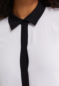 White shirt with a black collar and front placket, featuring smooth texture and simple design. Black buttons at the collar.