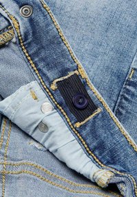 Denimbyxor med ljusblått tyg, gula sömmar, en mörk knapp och metallknappar, med en strukturerad yta och fransade kanter.