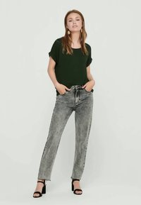 Frau steht mit den Händen in den Taschen, trägt ein schwarzes kurzärmliges Oberteil, graue Jeans im Acid-Wash-Stil, schwarze Sandalen mit Absatz und goldene Ohrringe.