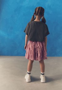 Graues Kurzarmoberteil, geschichteter pinkfarbener Rüschensrock, weiße Sneakers mit schwarzen Streifen auf Socken, von hinten vor einem blauen Hintergrund betrachtet.