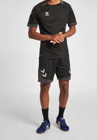 Camiseta deportiva negra de manga corta con acentos grises, combinada con pantalones cortos negros que presentan un patrón en forma de Chevron plateado, y zapatillas deportivas azules.