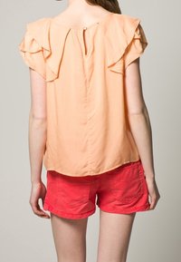 Persikofärgad blus med volangärmar, nyckelhål baktill med knappstängning, kombinerad med klara röda shorts med bakfickor.