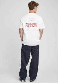 T-shirt blanche avec un texte rouge vif au dos : « LE SENTIMENT D'AMOUR DISPONIBLE POUR UN RENDEZ-VOUS. » Associée à un jean bleu foncé.