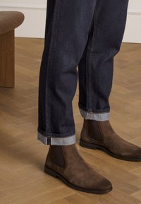 Personne portant un jean foncé à revers et des bottes Chelsea en suède marron debout sur un parquet en bois.