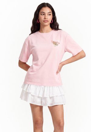 Kobieta ubrana w różowy t-shirt z małą grafiką i tekstem, połączony z białą, warstwową spódnicą z falbankami i różowymi kolczykami-kołami.