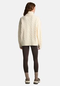 Ivory sweter w splot kablowy z wysokim kołnierzem, długimi rękawami i prążkowanym dołem, w połączeniu z brązowymi legginsami i taupe skarpetkami, na jednolitym tle.