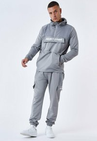 Grå træningsdragt med hætte, frontlomme og logo, lynlåsbukser med cargo-lommer og hvide sneakers. Glat, sporty stof.
