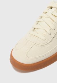Sapatilha de lona creme com um superior texturizado, fecho de atacadores tradicional e uma sola de borracha castanha em contraste.