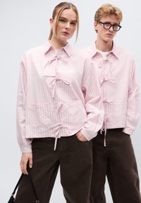 Παλ ροζ, υφασμάτινο σακάκι με κουμπιά και γιακά, κλείσιμο με κορδόνια και δύο μπροστινές τσέπες, φορεμένο με σκούρο καφέ παντελόνι.