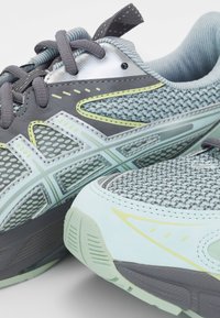 Gros plan de chaussures de running gris clair et vert clair avec des empeignes en mesh texturé et des surpiqûres synthétiques à motifs, montrant les lacets et les semelles.