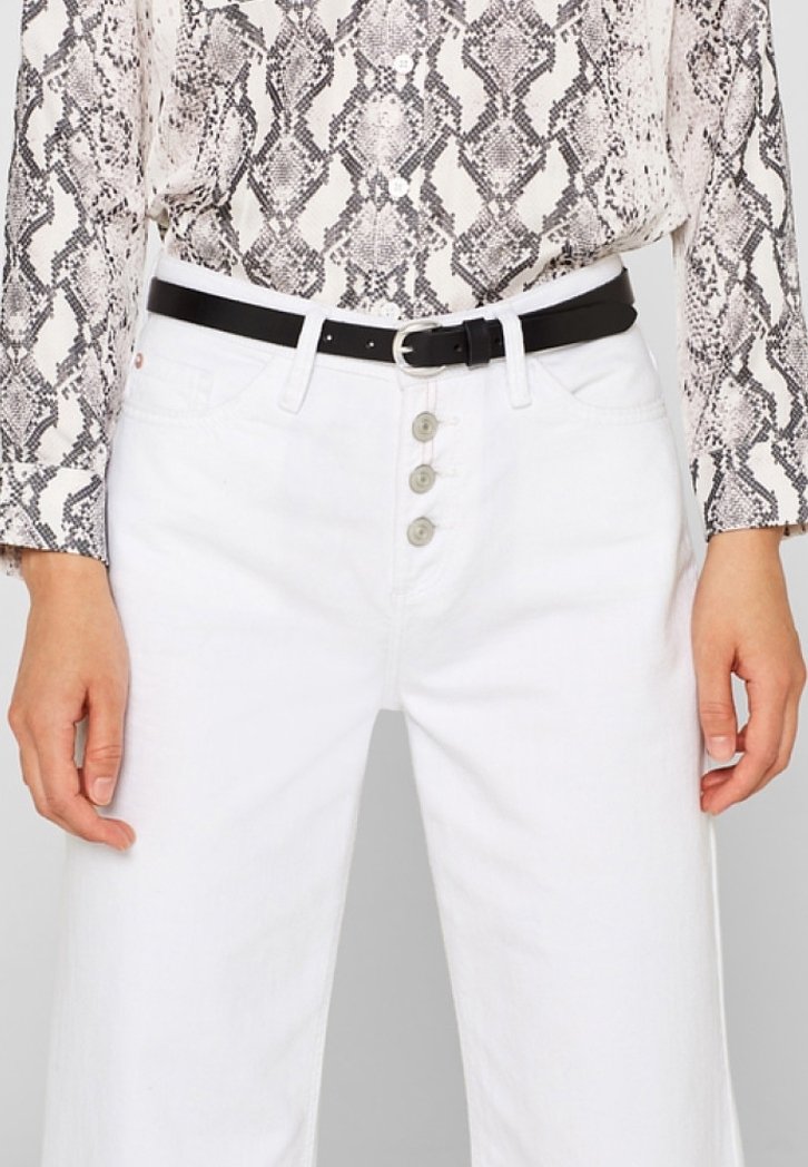 Personne portant un pantalon blanc à boutons avec une ceinture noire et une chemise à manches longues imprimé serpent, mains détendues le long du corps.