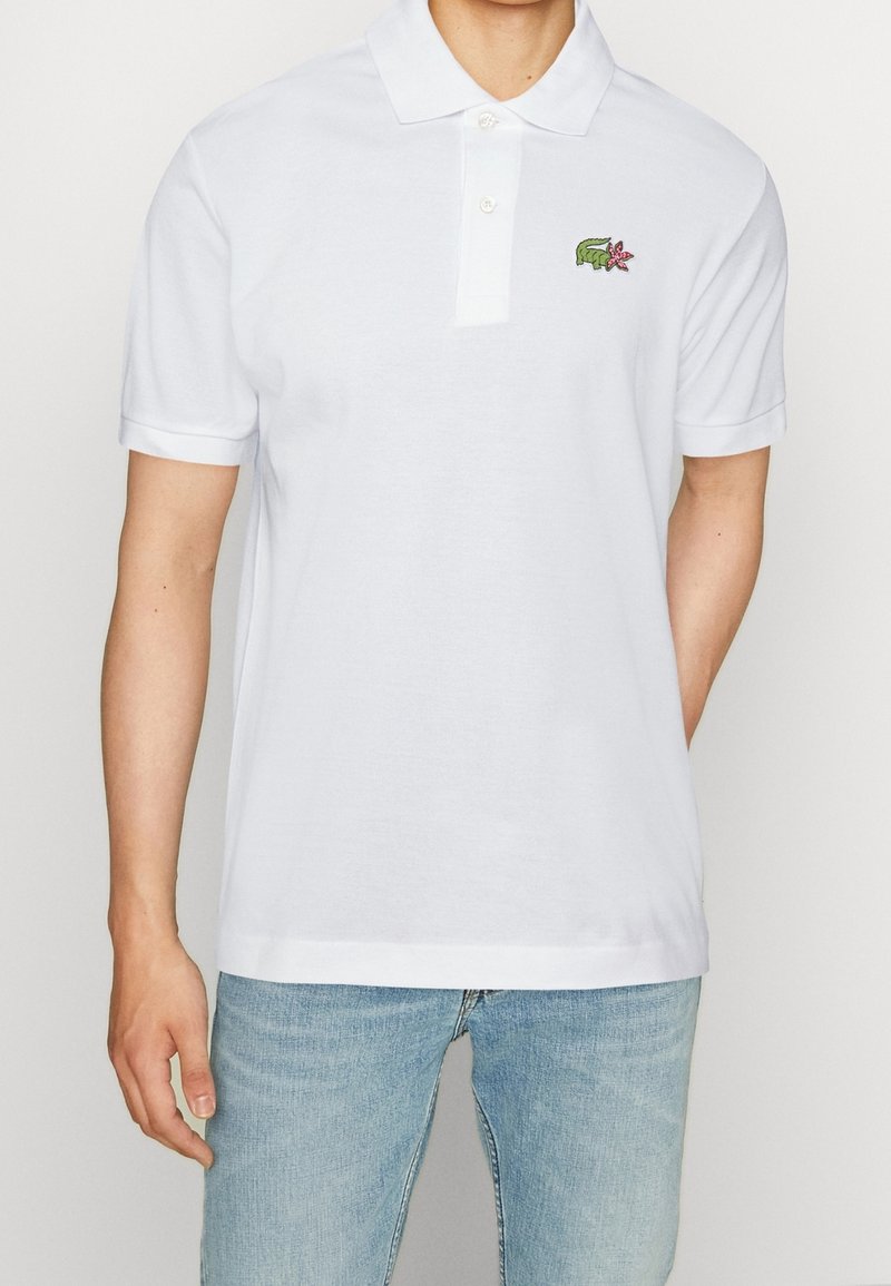 Polo shirt blanc à manches courtes, patte de boutonnage à deux boutons, et logo de crocodile vert brodé avec accent de fleur rose sur le côté gauche de la poitrine.