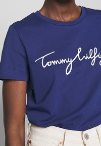 Námořnická modrá bavlněná tričko s velkým bílým kurzívním logem. Kulatý výstřih, krátké rukávy a hladká textura. Nosené s světlými kalhotami.