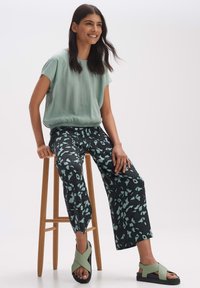 Mintgroene top met korte mouwen, zwarte broek met een abstract lichtgroen patroon en groene sandalen met kruisbandjes.