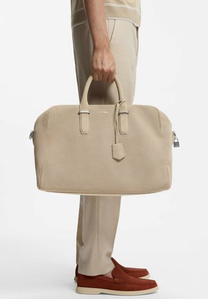 Person holding a large beige perforated leather duffel bag with handles, wearing beige pants and brown loafers, against a plain background.