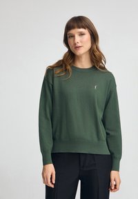 Pull en maille verte avec un col rond et des poignets côtelés. Présente un petit accent brodé sur la poitrine. Associé à un pantalon noir.