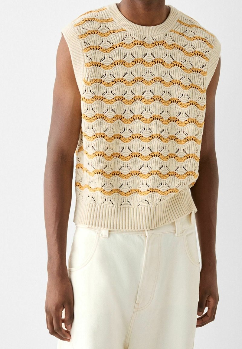 Top senza maniche color crema in maglia con motivi a onda in beige e oro, con orlo a costine. Indossato sopra pantaloni chiari.