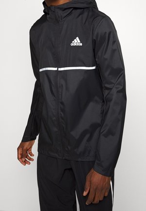 Mężczyzna ubrany w czarną kurtkę z kapturem Adidas z białym logo i poziomym paskiem, w połączeniu z czarnymi spodniami na jednolitym tle.