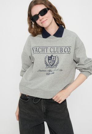 Młoda kobieta w czarnych okularach przeciwsłonecznych, szarej bluzie z emblematem "Yacht Club Co.", granatowym kołnierzem i czarnych dżinsach, pozująca z ręką na biodrze.