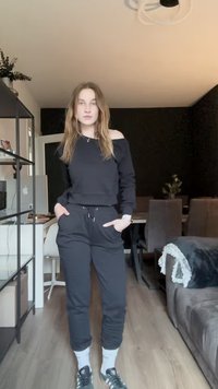 Sweatshirt preto com design de ombro à mostra, combinado com calças de jogging pretas. Apresenta punhos e cintura elásticos, usado com meias brancas e sapatos escuros.