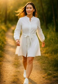 Abito bianco con bottoni, maniche lunghe e vita legata, realizzato in tessuto leggero, abbinato a sneakers bianche, che cammina su un sentiero.