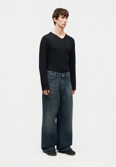 Jeune homme debout, portant une chemise noire à manches longues et un jean large bleu délavé oversized avec des chaussures noires, sur un fond uni.