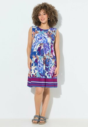 Femme aux cheveux bouclés portant une robe sans manches à fleurs dans des tons bleus, violets et roses, debout contre un mur blanc, chaussée de sandales bleues.