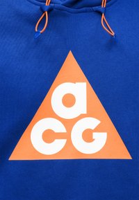 Μπλε φούτερ με κουκούλα με πορτοκαλί τρίγωνο που έχει λευκά γράμματα "a," "c," και "g" μέσα· πορτοκαλί κορδονάκια με μπλε άκρα.