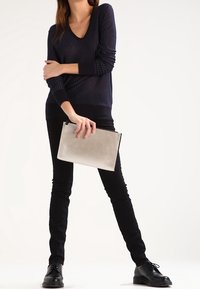 Suéter de punto canalé en color azul marino con escote en V, pantalones skinny negros y zapatos de cordones negros. Sosteniendo un bolso clutch rectangular en color gris claro.