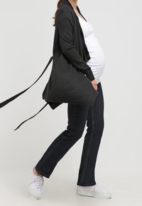 Svart cardigan med draperad front och öppen design, som matchas med en vit linne och mörka denimjeans. Modellen har en rund mage och bär vita sneakers.