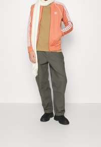 Herrkläder med en orange zip-up jacka med vita ränder, brun t-shirt, grå cargobyxor och svarta skor, kompletterat med en krämfärgad halsduk.