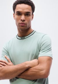 Ανοιχτοπράσινο κοντομάνικο μπλουζάκι με γιακά με μαύρες λεπτομέρειες και λογότυπο "BOSS" στον λαιμό, φορεμένο από άτομο με σταυρωμένα χέρια.