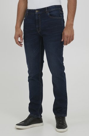 Mørkeblå jeans i slim fit med fem lommer, avsluttet med gult søm og knappeglidelås. Brukt med svarte sneakers.