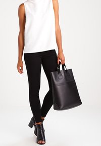 Bolso tote de cuero negro con dos asas cortas, forma rectangular, textura suave y una línea de acento horizontal. Modelo con una blusa sin mangas blanca y leggings negros.