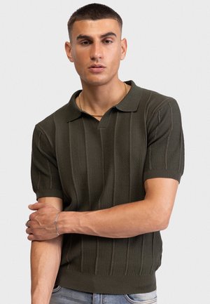 Άνδρας με κοντά μαλλιά που φοράει ένα σκούρο πράσινο πλεκτό κοντομάνικο πόλο, χρυσό αλυσίδα στο λαιμό, ασημένιο βραχιόλι και ανοιχτό μπλε τζιν.