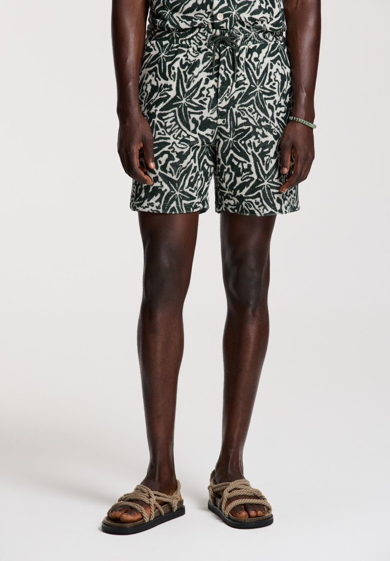 Eine schwarzhäutige Person trägt grün-weiße Shorts mit Sternmuschel-Muster und beigen Seil-Sandalen und steht vor einem schlichten weißen Hintergrund.