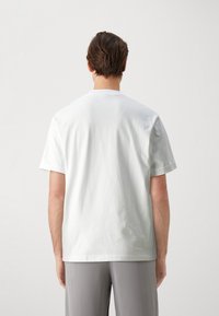 Bílý bavlněný tričko s krátkými rukávy, kulatým výstřihem a volným střihem, zobrazený zezadu. Hladký povrch, minimalistický design.