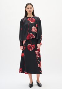 Donna in piedi, che indossa un abbinato top nero e una gonna midi con grande stampa floreale rossa e rosa, e scarpe nere con punta, su sfondo bianco.