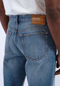 Jeans in denim azzurro chiaro con una vestibilità aderente, dotati di cinque tasche e cuciture arancioni a contrasto. L'etichetta in pelle sulla vita reca la scritta "DRDENIM."